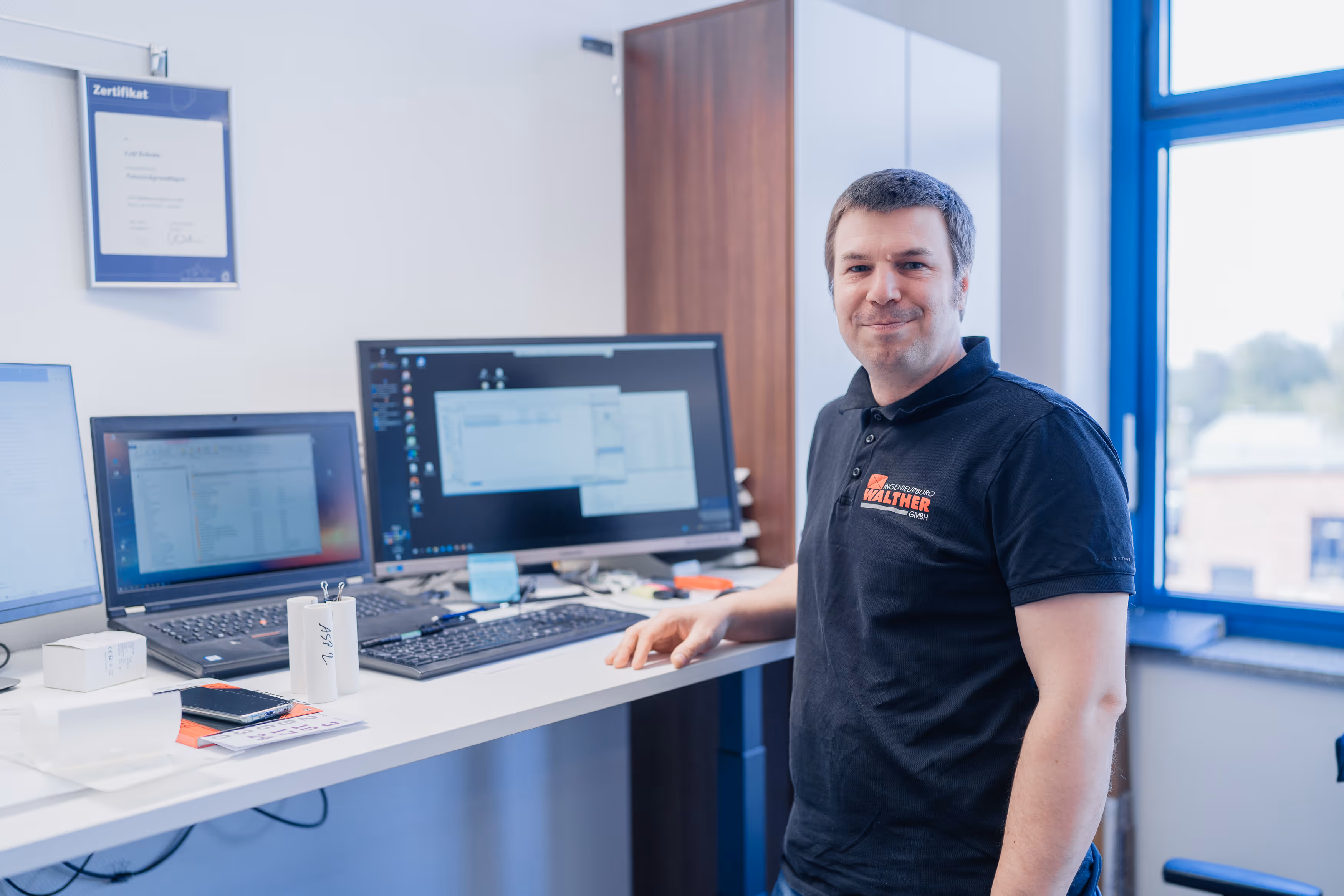 Mann in schwarzem Poloshirt mit Walther-Logo steht an einem Schreibtisch mit drei Bildschirmen in einem Büro mit Fenster.