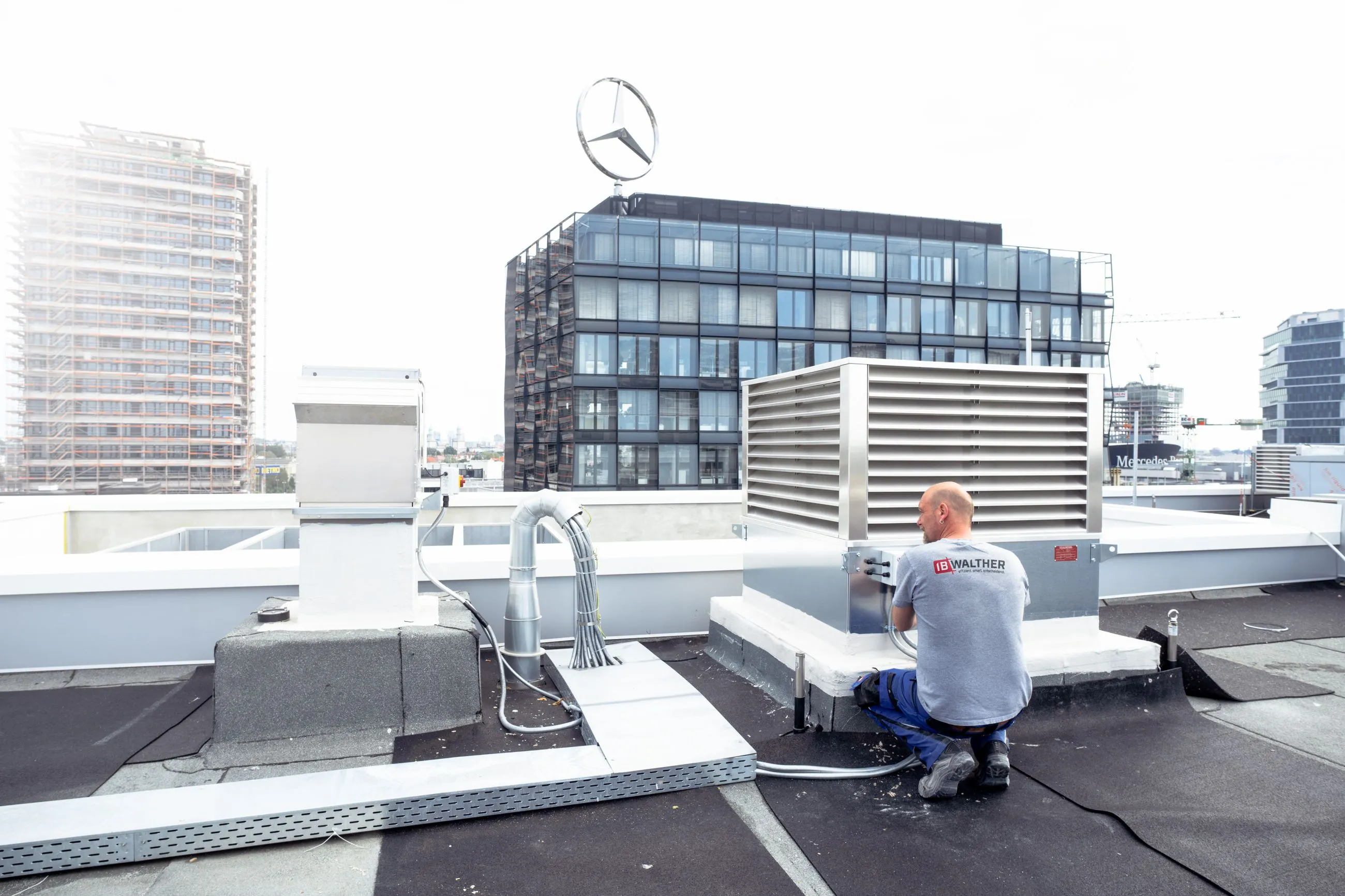 Techniker von IB Walther in Arbeitskleidung arbeitet an einer Belüftungsanlage auf dem Dach eines Gebäudes mit Mercedes-Stern im Hintergrund.