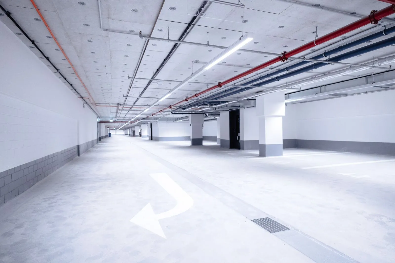 Helle, leere Tiefgarage mit weißen Wänden, Pfeilen auf dem Boden und sichtbaren Rohrleitungen an der Decke.