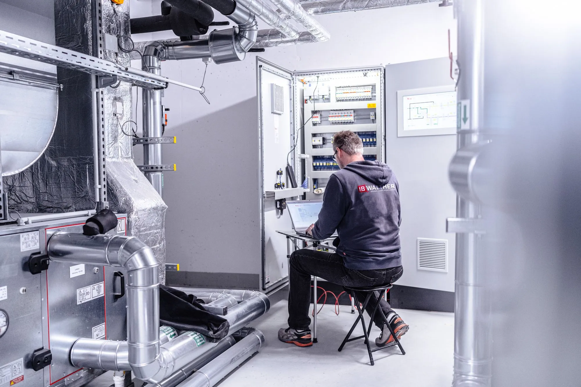 Techniker arbeitet an Laptop vor offenem Schaltschrank in einem Raum mit Lüftungsrohren.