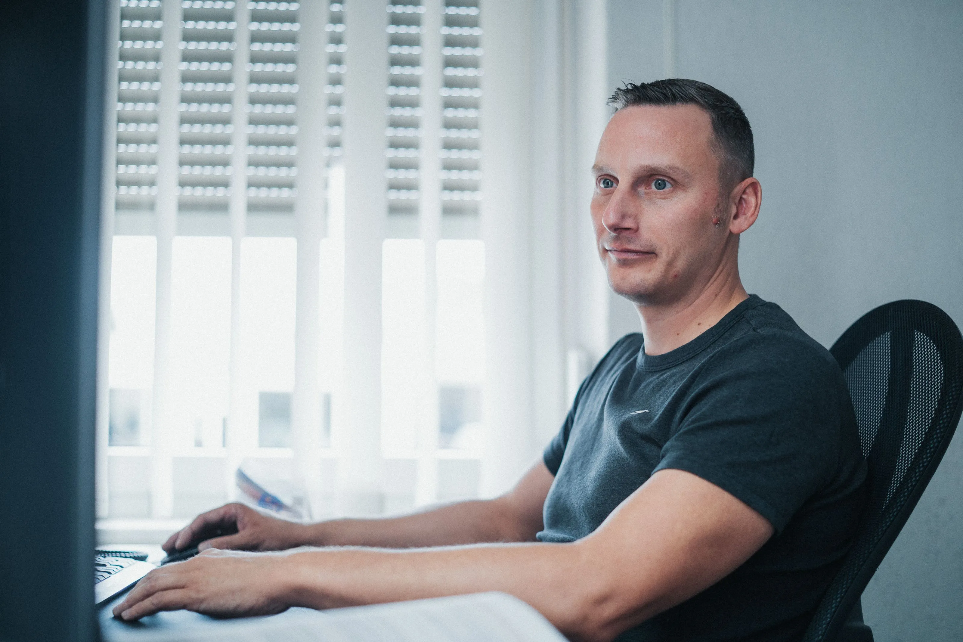 Mann mit schwarzem T-Shirt sitzt an einem Schreibtisch und arbeitet an einem Computer vor einem Fenster mit vertikalen Jalousien.