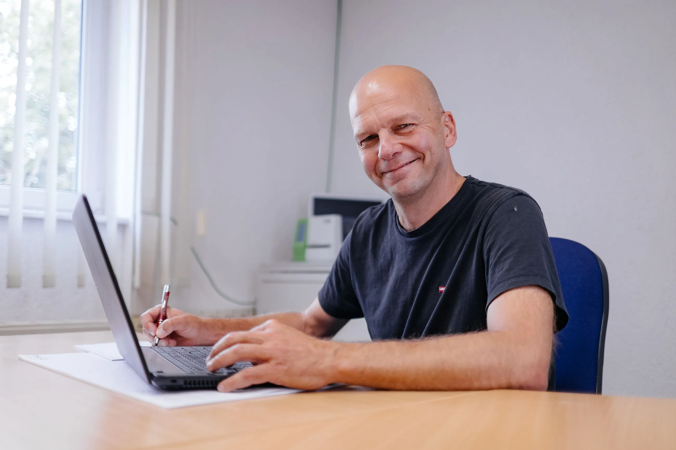 Ein Mann in schwarzem IB Walther T-Shirt sitzt an einem Schreibtisch und arbeitet an einem Laptop, während er lächelt.