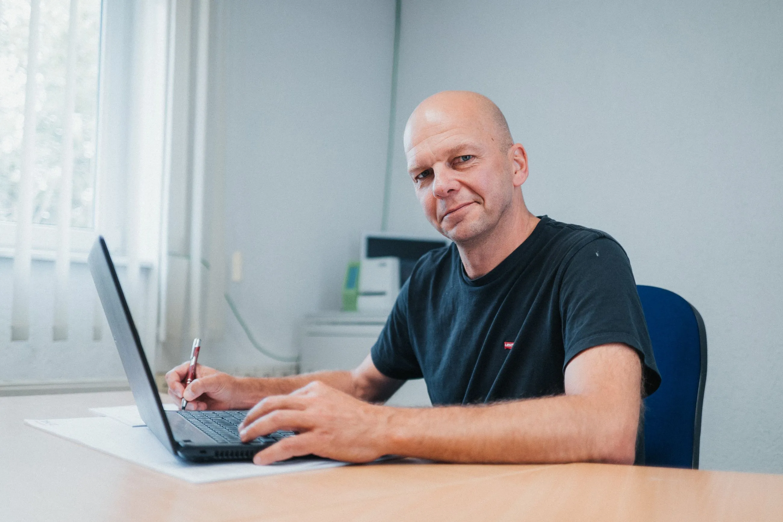 Mann in IB Walther T-Shirt sitzt an einem Schreibtisch, tippt auf einem Laptop und hält einen Stift.