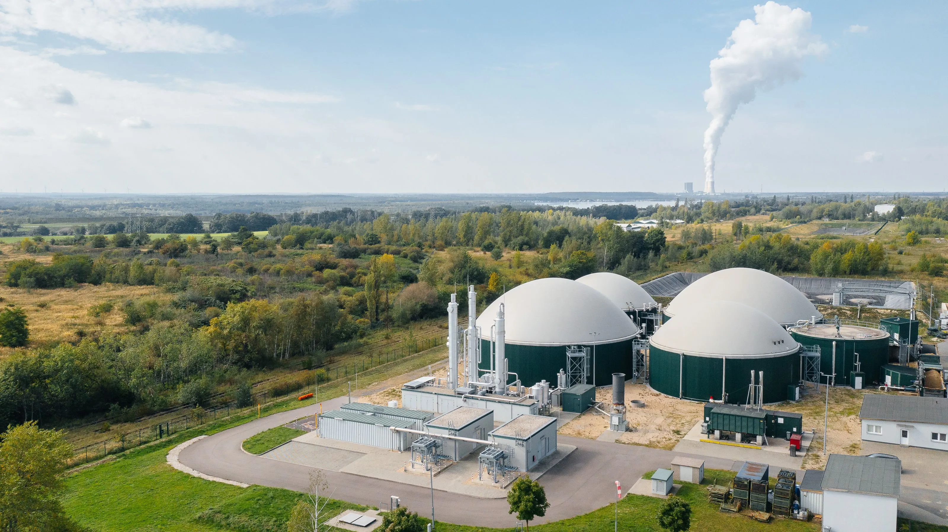 Luftaufnahme einer Biogasanlage mit vier kuppelförmigen Fermentern in einer grünen, ländlichen Umgebung unter blauem Himmel.