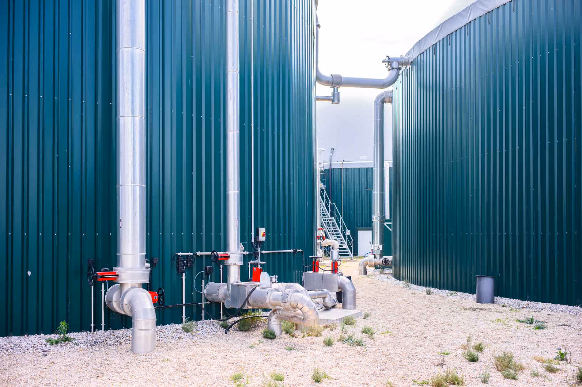 Große grüne Industriebehälter mit silbernen Rohrleitungen und Ventilen auf steinigem Boden.