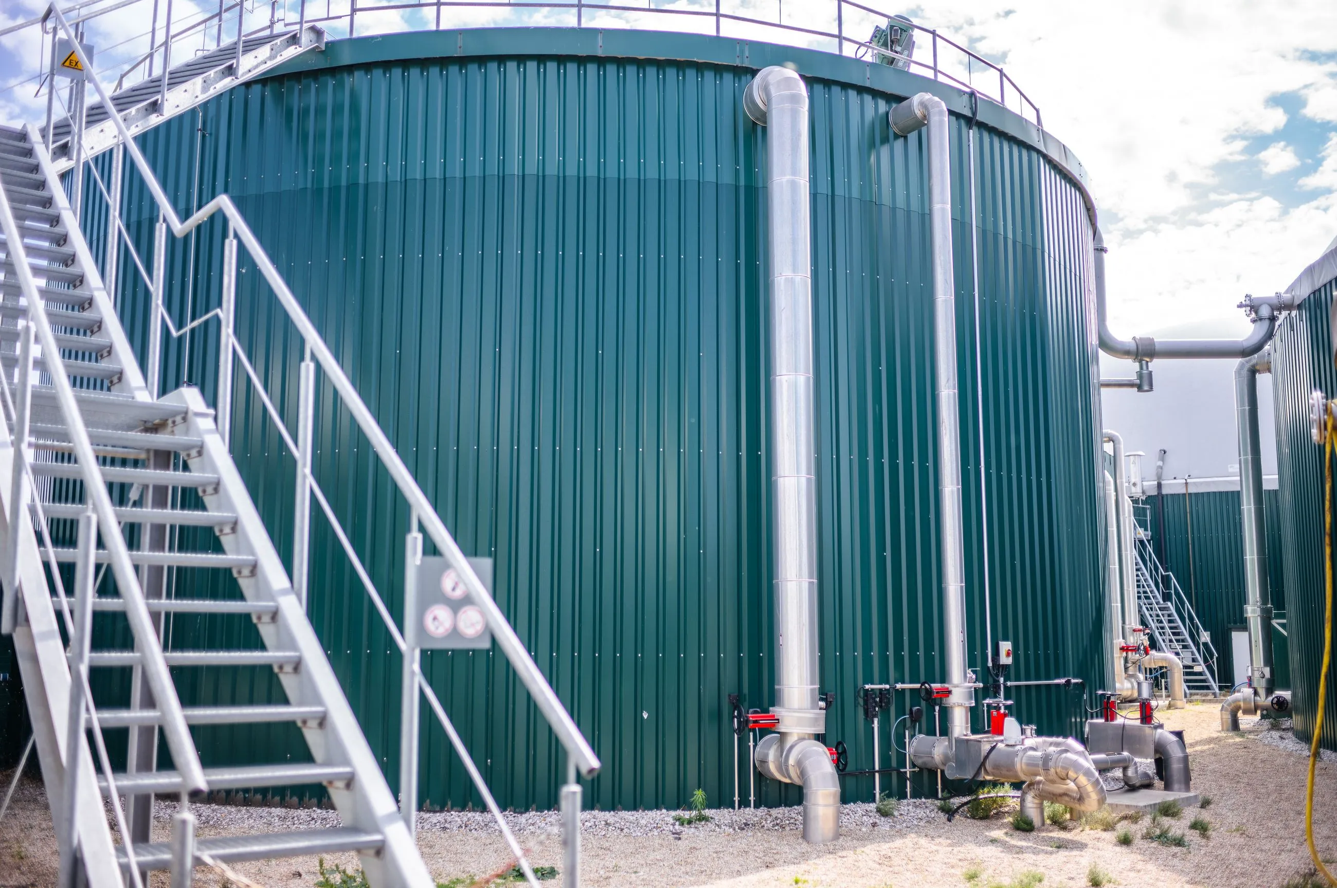 Großer grüner Industrietank mit Metallrohren und einer Metalltreppe im Freien unter bewölktem Himmel.