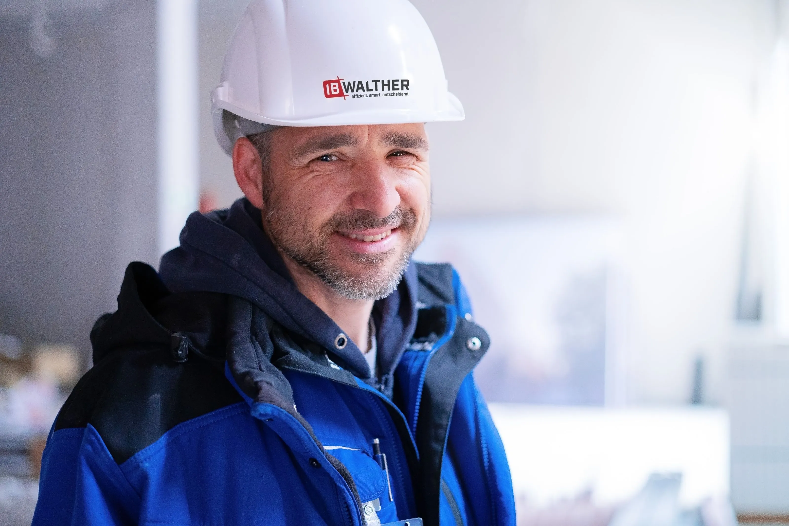 Lächelnder Bauarbeiter mit weißem Helm und blauer Arbeitsjacke in einer hellen Innenumgebung.