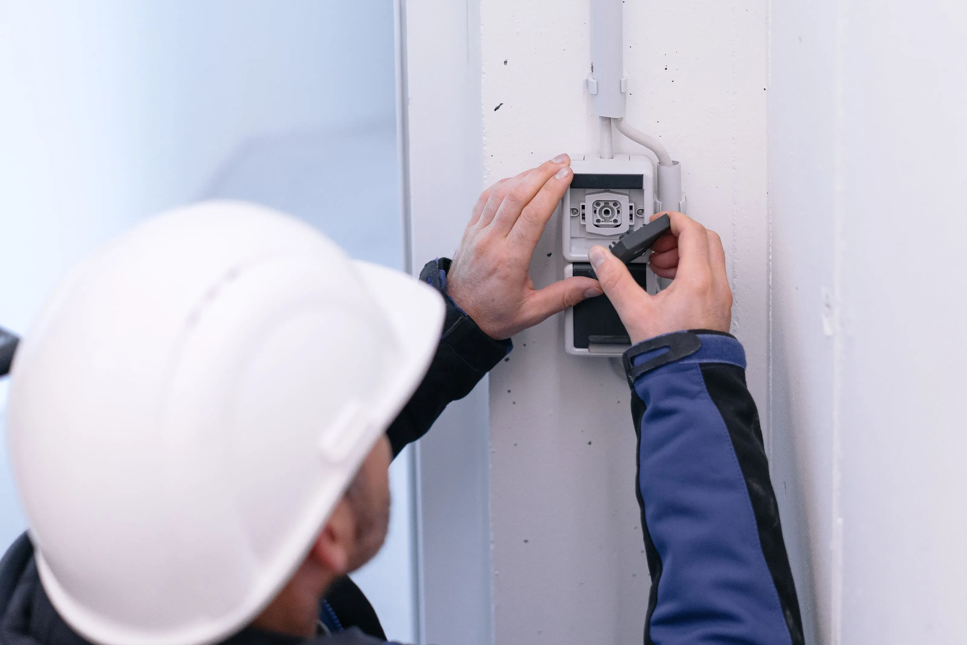 Elektriker mit weißem Helm montiert Außensteckdose an Wand.