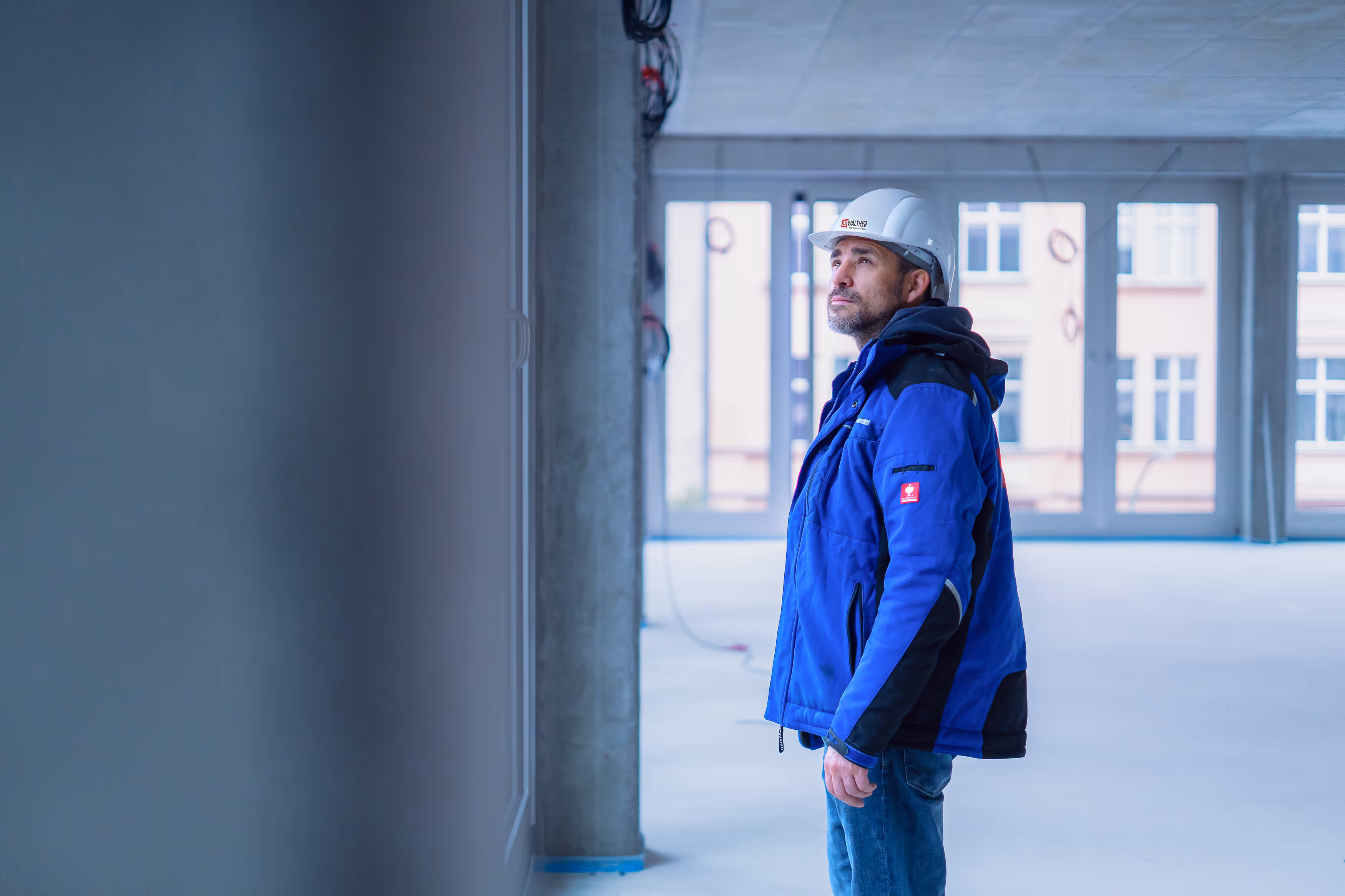 Bauarbeiter mit weißem Helm und blauer Jacke inspiziert Innenraum einer Baustelle mit großen Fenstern.