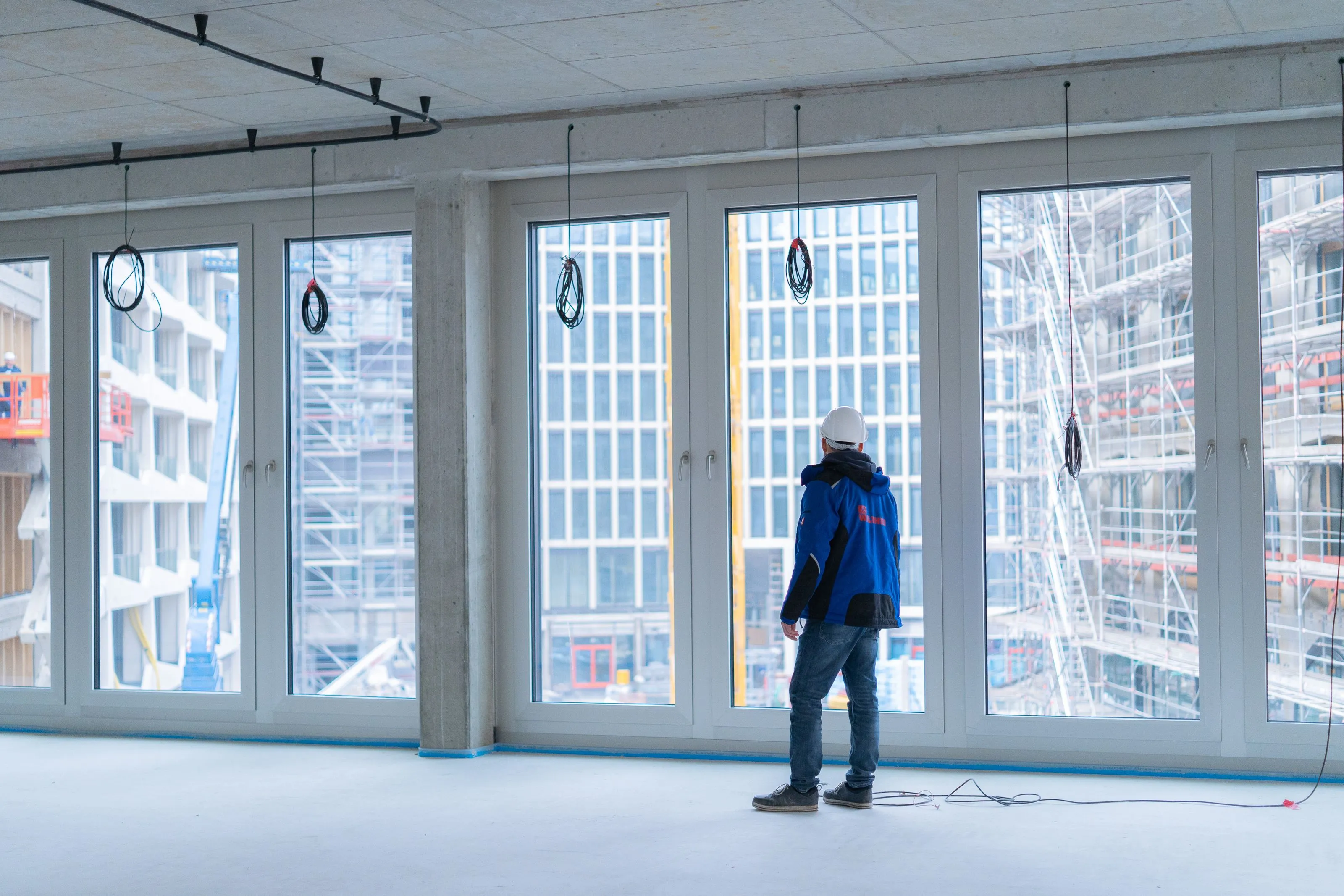 Bauarbeiter mit Helm und blauer Jacke steht in einem unfertigen Raum und blickt aus großen Fenstern auf eine Baustelle mit Gerüsten.