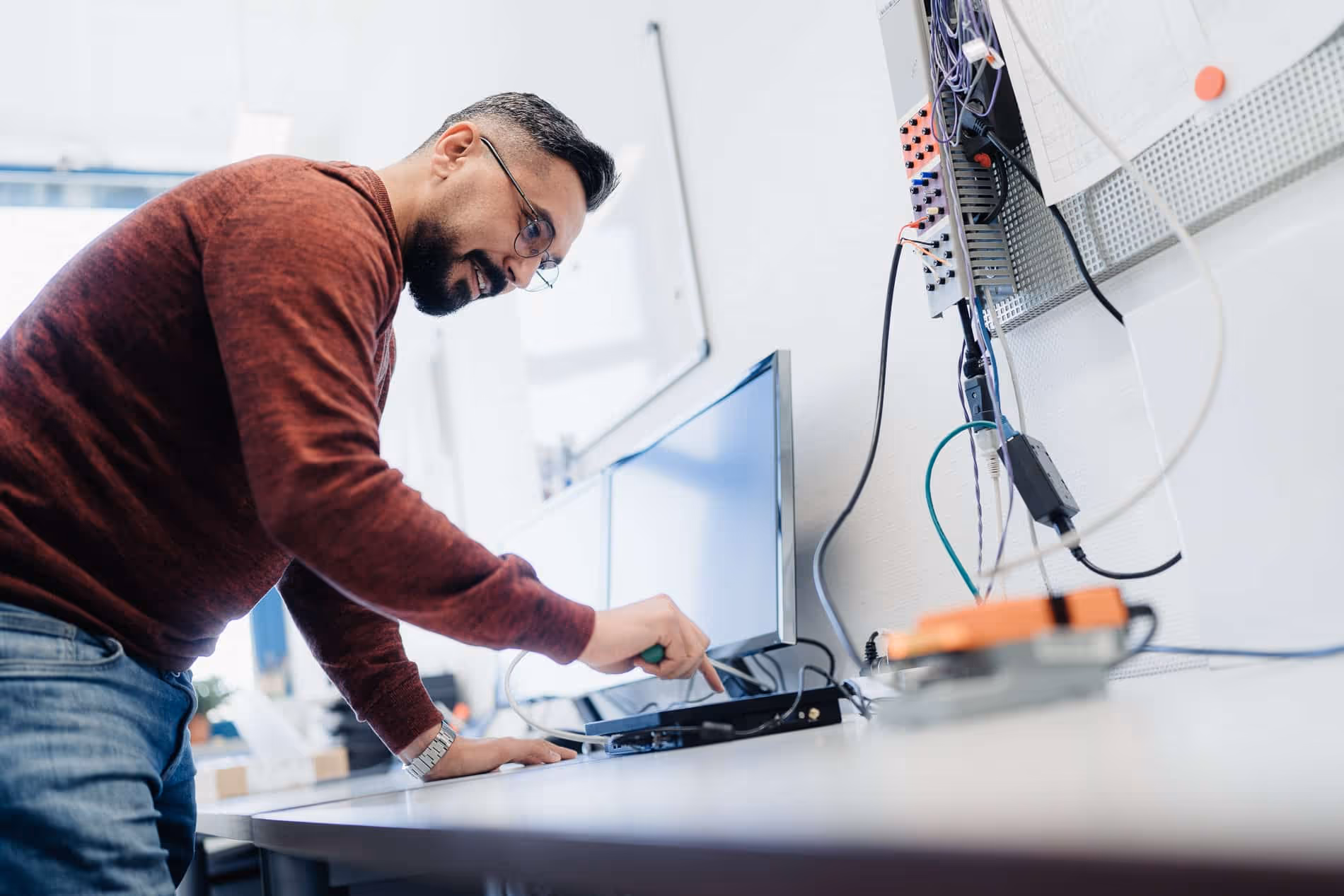Mann mit Brille und rotem Pullover arbeitet an einem technischen Gerät an einem Schreibtisch mit Computermonitor und verkabelten Steuerungen.