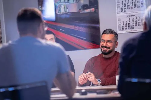 Mann mit Brille und rotem Pullover lächelt in einer Besprechung mit mehreren Personen um einen Tisch.