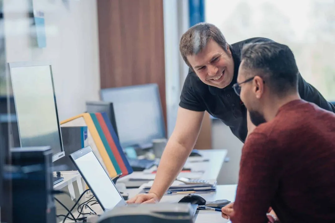 Zwei Männer unterhalten sich lachend in einem Büro vor einem Laptop.