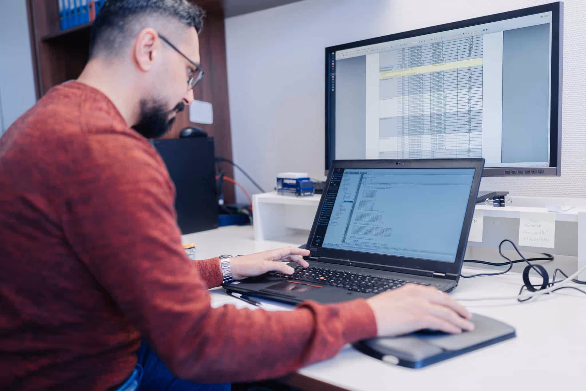 Ein Mann mit rotem Pullover arbeitet an einem Laptop vor einem großen Monitor mit einer Tabellenkalkulation.