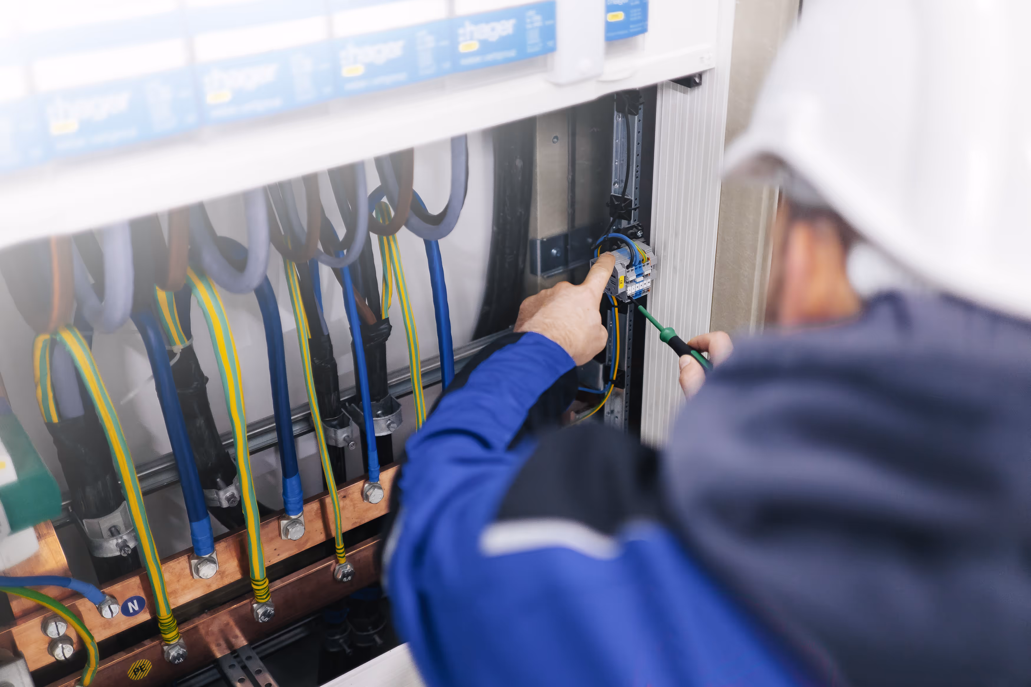 Elektriker in blauem Arbeitsmantel und weißem Helm arbeitet an einem Sicherungskasten mit Schraubendreher.