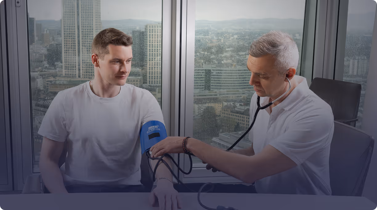 Healthcare professional measuring a young man's blood pressure in an office with large windows overlooking a cityscape on a rainy day.
