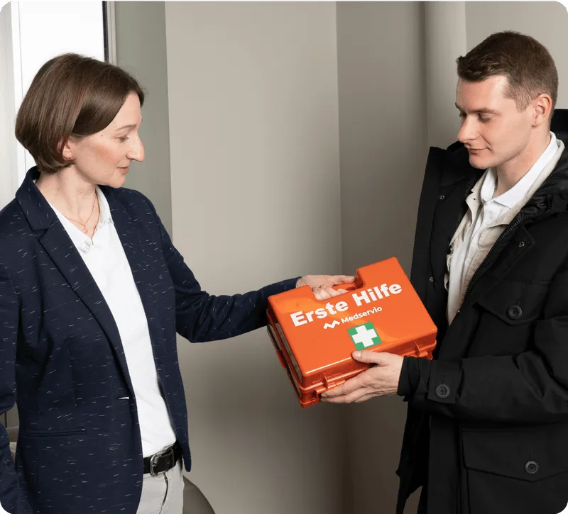 A woman handing an orange Erste Hilfe first aid kit to a man indoors.