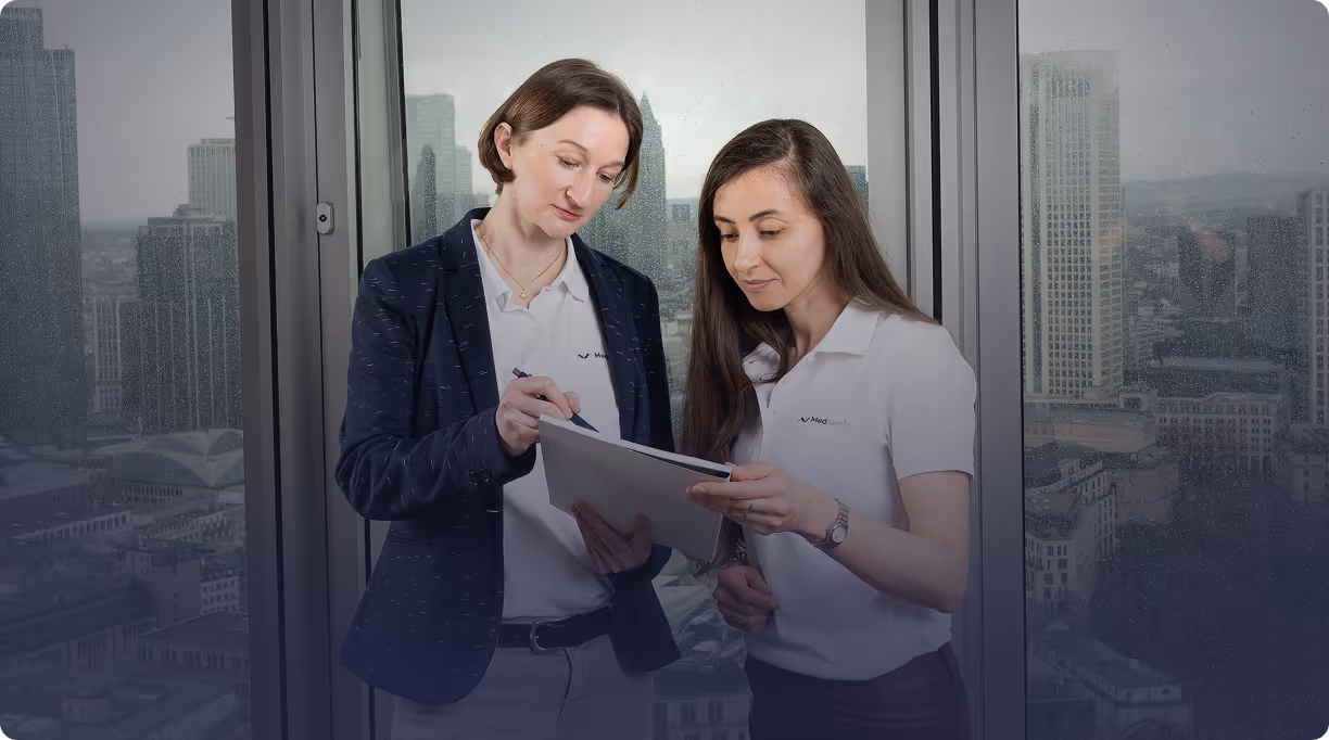 Two factory workers wearing white hard hats and yellow safety vests, standing and holding a tablet in an industrial setting.