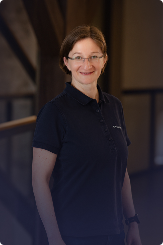 Eine lächelnde Frau mit Brille und kurzen braunen Haaren trägt ein dunkles Poloshirt und steht vor einem unscharfen, dunklen Hintergrund.