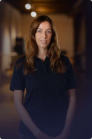 Porträt einer lächelnden Frau mit langen braunen Haaren, die ein schwarzes Poloshirt trägt, vor einem unscharfen, warm beleuchteten Hintergrund.