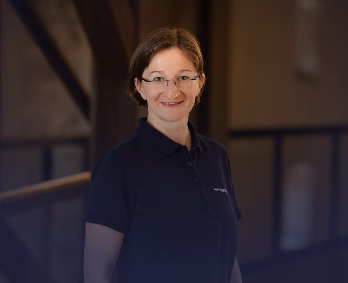 Lächelnde Frau mit kurzen braunen Haaren und Brille, trägt ein dunkelblaues Poloshirt vor unscharfem Hintergrund.