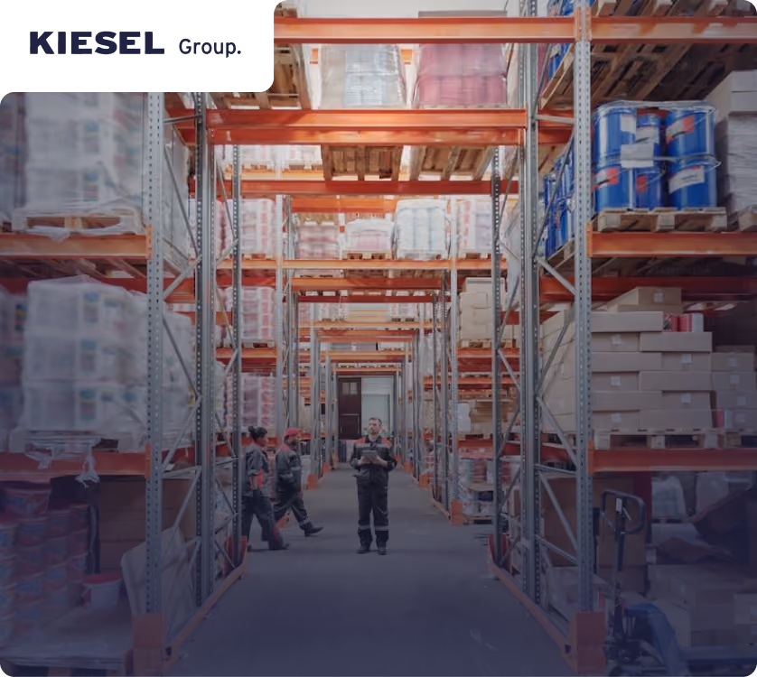 Warehouse aisle with tall shelves stocked with boxes and containers, and four workers standing and walking along the aisle.