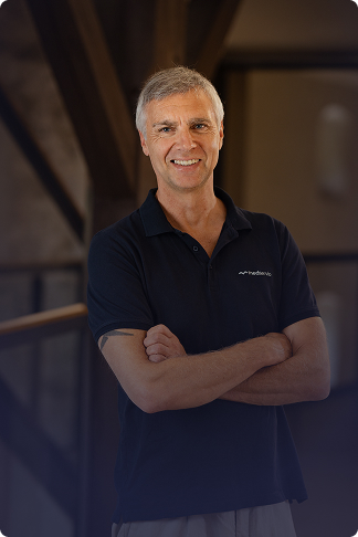 Ein lächelnder Mann mittleren Alters mit grauem Haar, der einen schwarzen Poloshirt mit Medservio-Logo trägt und die Arme vor der Brust verschränkt hat.