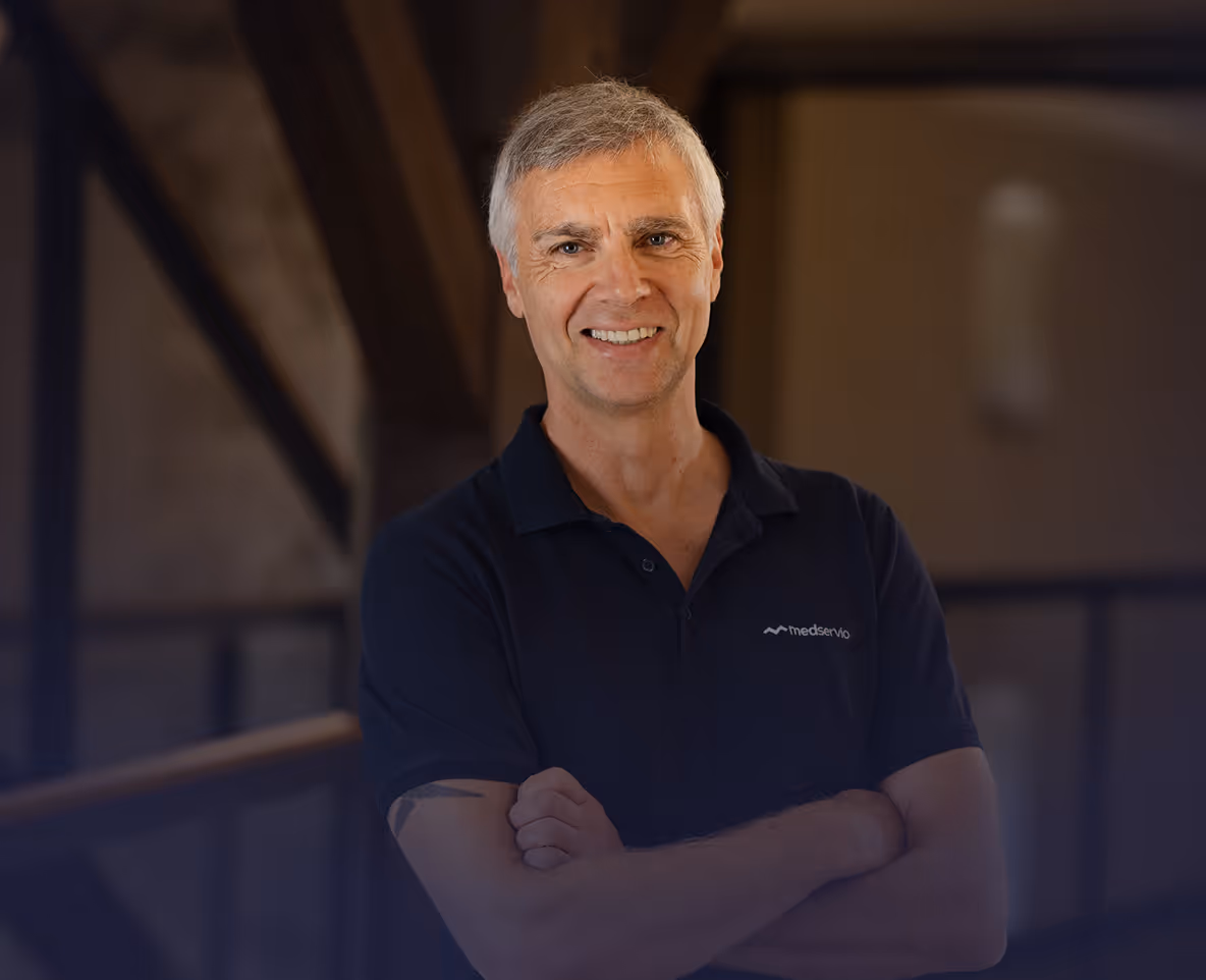 Lächelnder Mann mit kurz grauem Haar, der verschränkt die Arme trägt und ein dunkles Medservio-Poloshirt trägt.