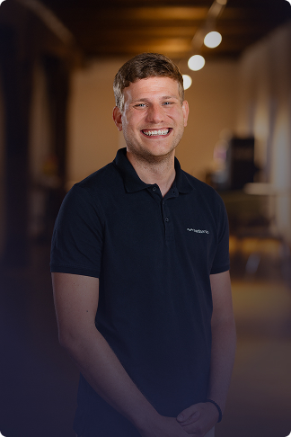 Lächelnder junger Mann mit kurzbraunen Haaren trägt dunkelblaues Poloshirt in einem warm beleuchteten Raum.