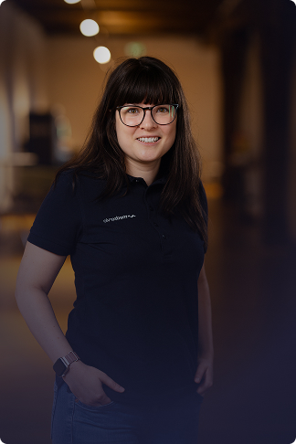 Frau mit Brille, dunkelbraunen Haaren und blauem Poloshirt lächelt vor unscharfem Hintergrund.