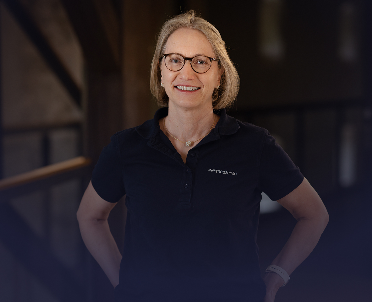 Lächelnde Frau mit Brille, blondem Haar und schwarzem Medservio-Poloshirt vor dunklem unscharfem Hintergrund.
