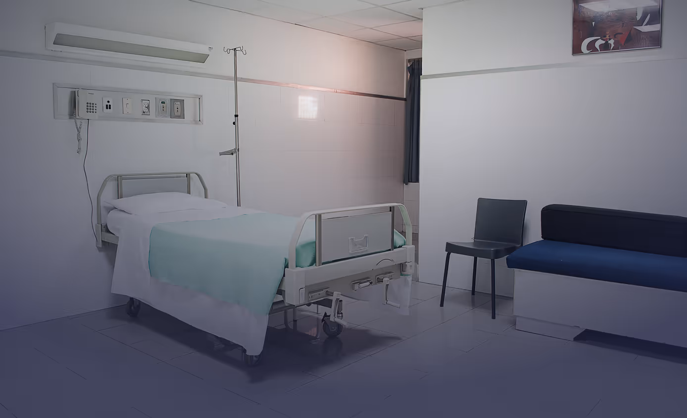 Krankenhauszimmer mit einem Bett, Infusionsständer, Stuhl und Sofa vor weißen Wänden.