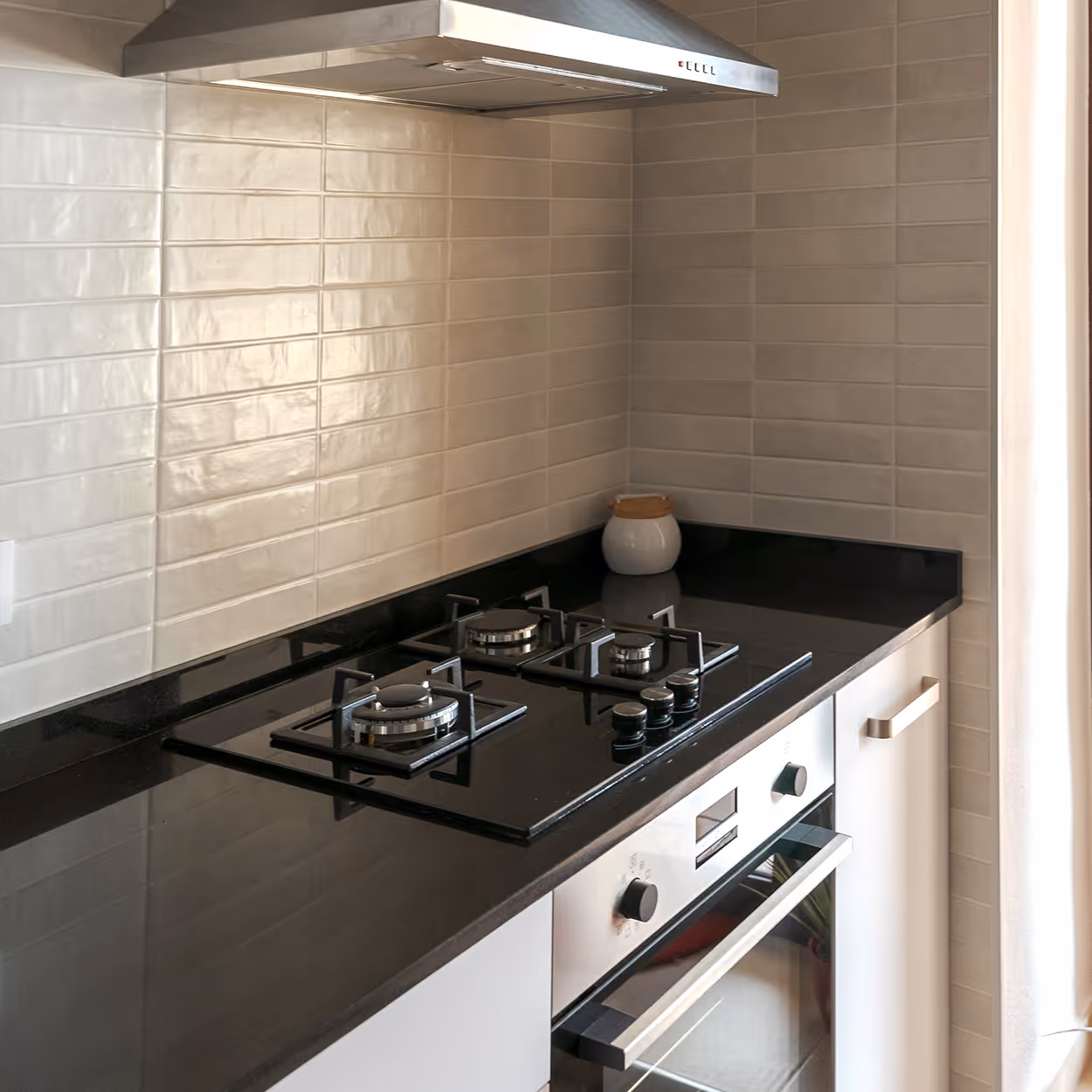 Modern kitchen corner with black gas stove, stainless steel oven, beige tiled backsplash, and a jar on the countertop.