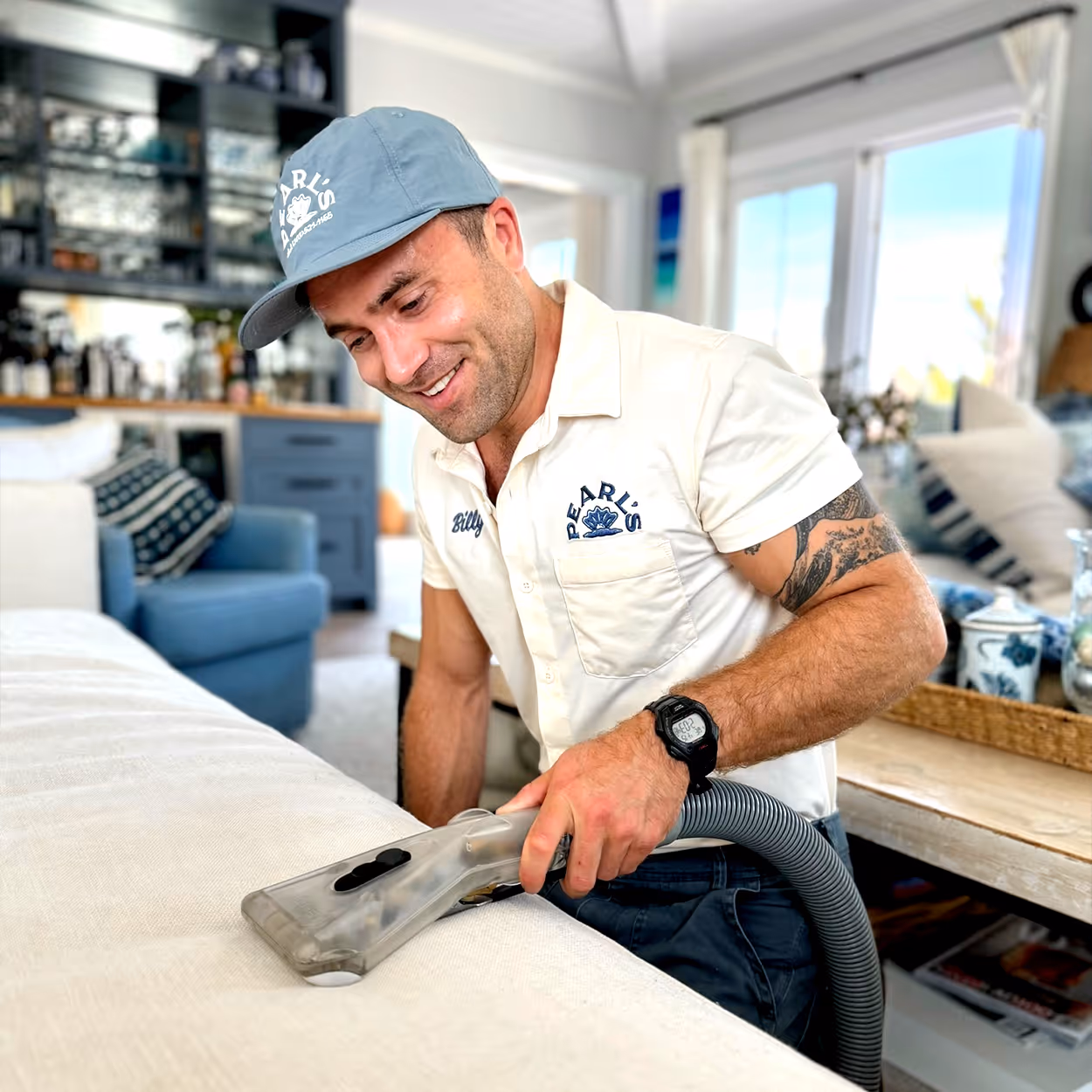 Man wearing a Pearl's uniform cleaning a white couch with a handheld upholstery vacuum in a bright living room.