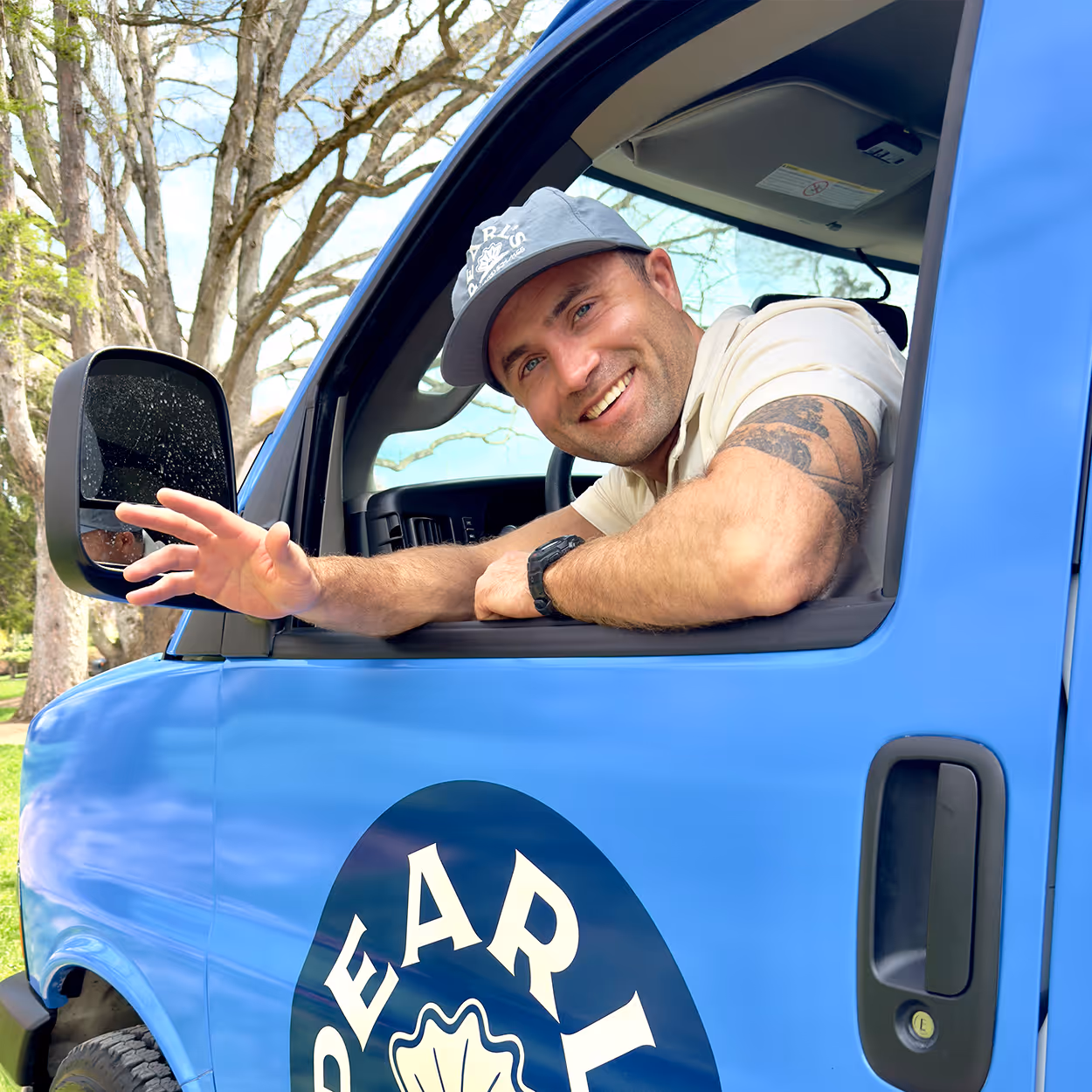 Smiling man wearing a gray cap waving from the driver's seat of a blue vehicle with a partial circular logo on the door.