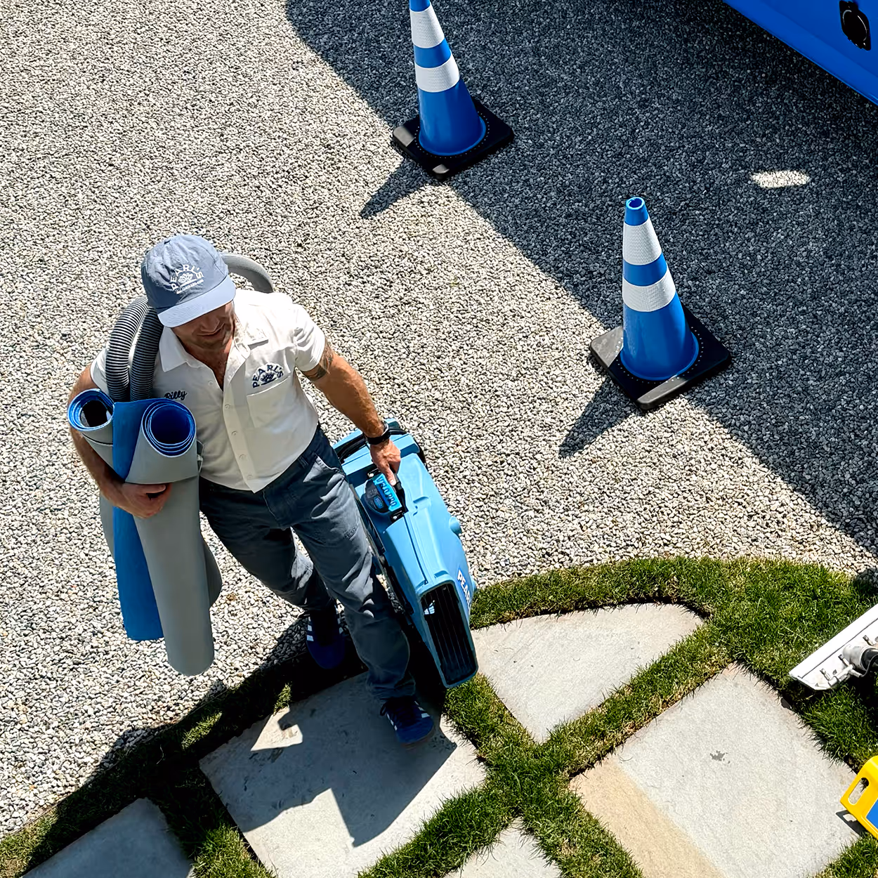 Man carrying rolled mats and a blue vacuum cleaner walking on a gravel and grass pathway near traffic cones.