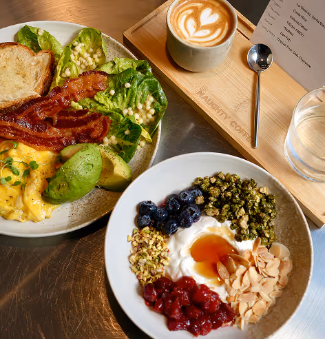 Breakfast spread with scrambled eggs, bacon, avocado, lettuce, toast, yogurt bowl with granola, berries, almonds, honey, and a cup of coffee with latte art on a wooden tray.