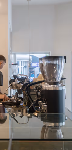 Close-up view of an espresso machine and coffee grinder on a reflective countertop with a barista working in the background.