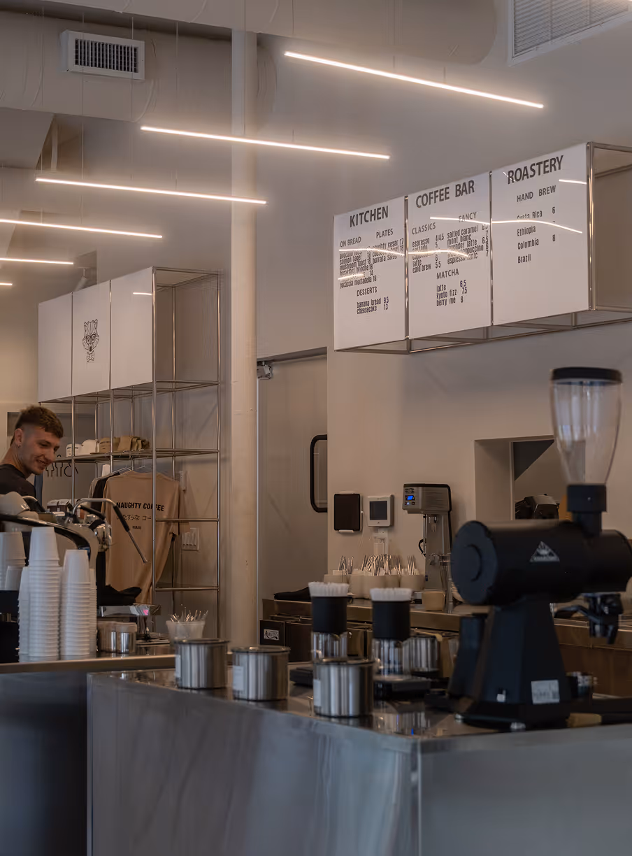 Modern coffee shop interior with a barista behind the counter, coffee machines, and illuminated menu boards for kitchen, coffee bar, and roastery options.