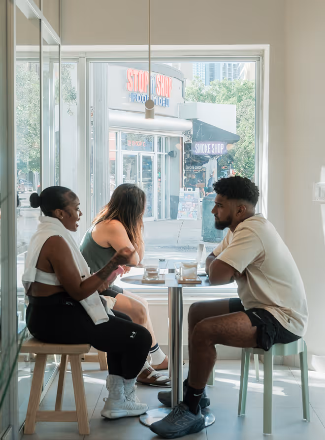 Three people sitting inside a cafe, chatting around a small table near a large window with a view of a street and storefronts outside.