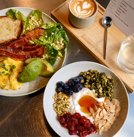 Breakfast plates with bacon, avocado, scrambled eggs, salad, yogurt bowl with fruit and granola, and a latte.