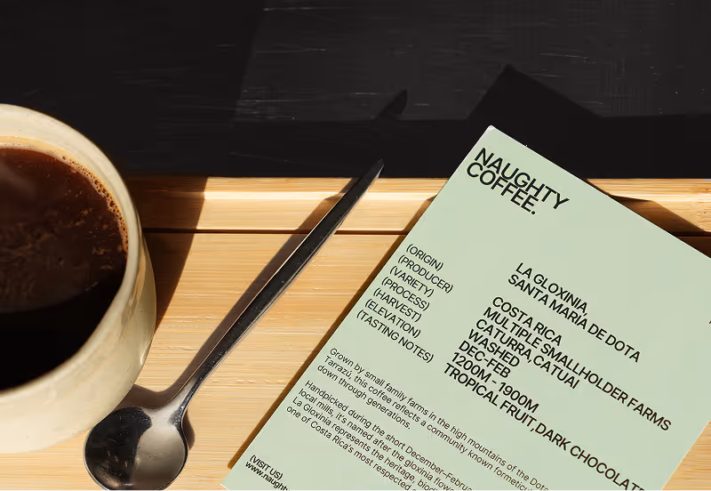 Cup of coffee beside a spoon and a card listing details of Naughty Coffee from Costa Rica.