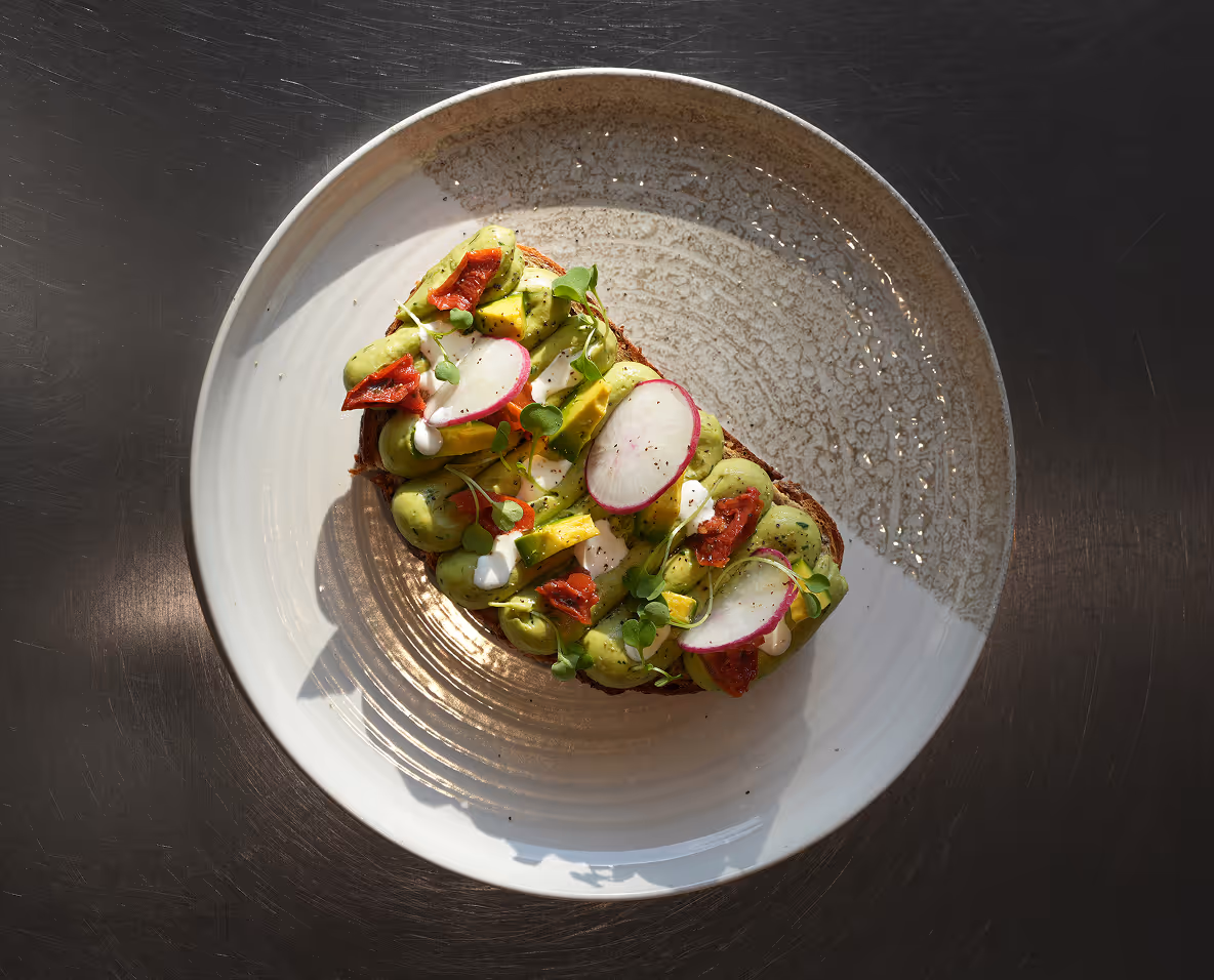 Toast topped with avocado, radish slices, sun-dried tomatoes, microgreens, and a drizzle of white sauce on a round plate.