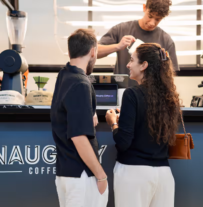 Two customers in black tops order coffee at a coffee shop counter with a barista pouring milk.