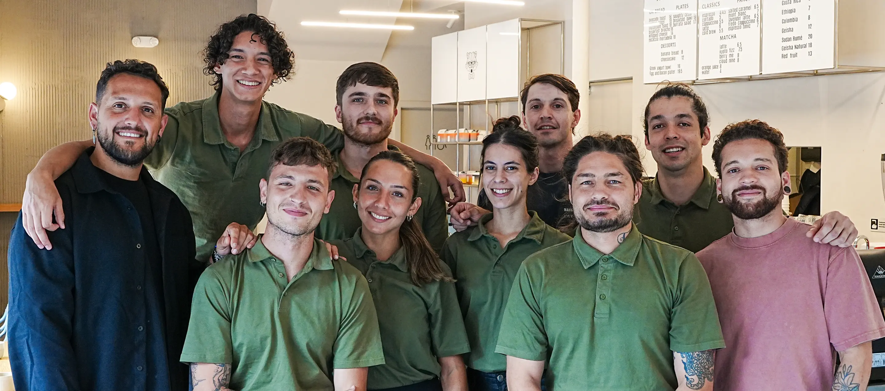 Group of ten smiling restaurant staff, most wearing green polo shirts, posing together indoors.