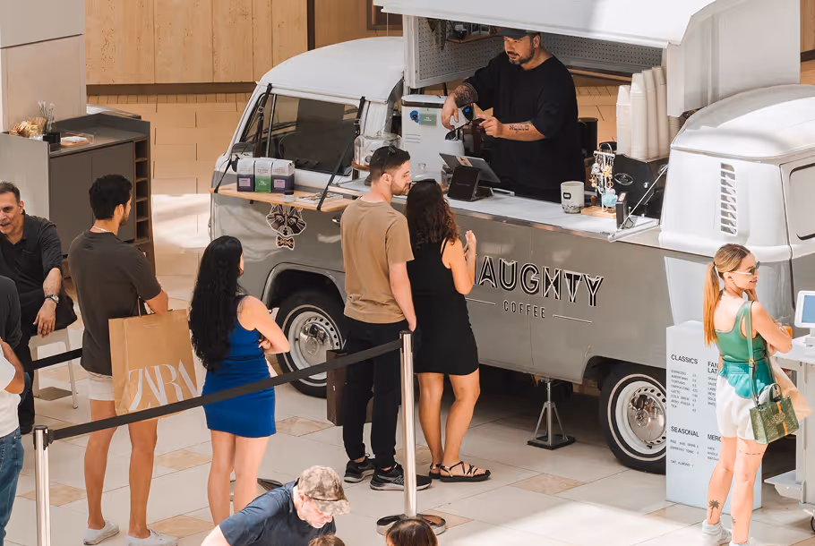 People wait in line at Naughty Coffee truck inside a mall while a barista prepares a drink.