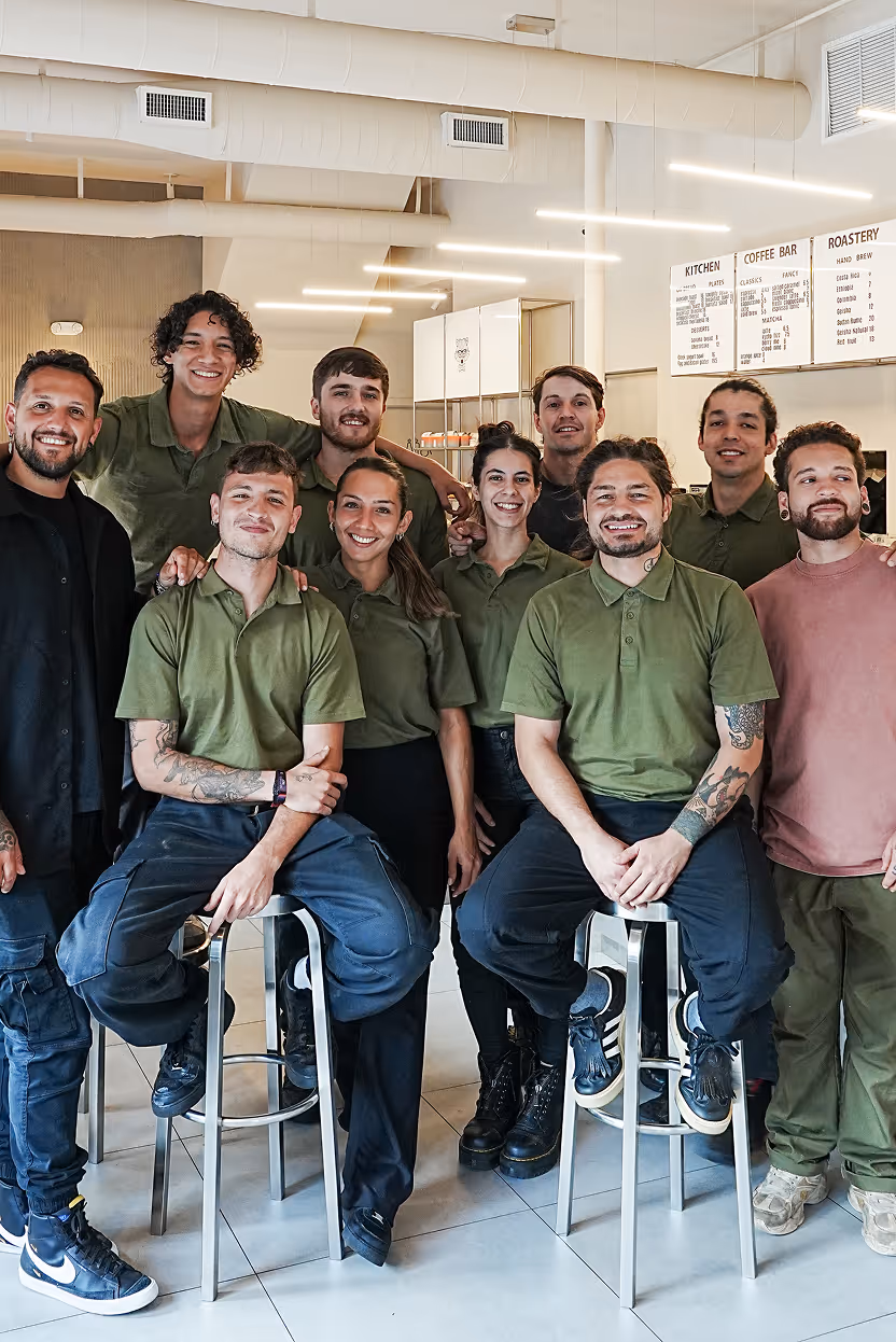 Group of smiling young café staff in green shirts posing together inside a modern coffee shop.
