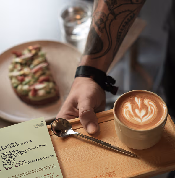 Hand holding wooden tray with latte art coffee cup, spoon, menu, and toast in background.