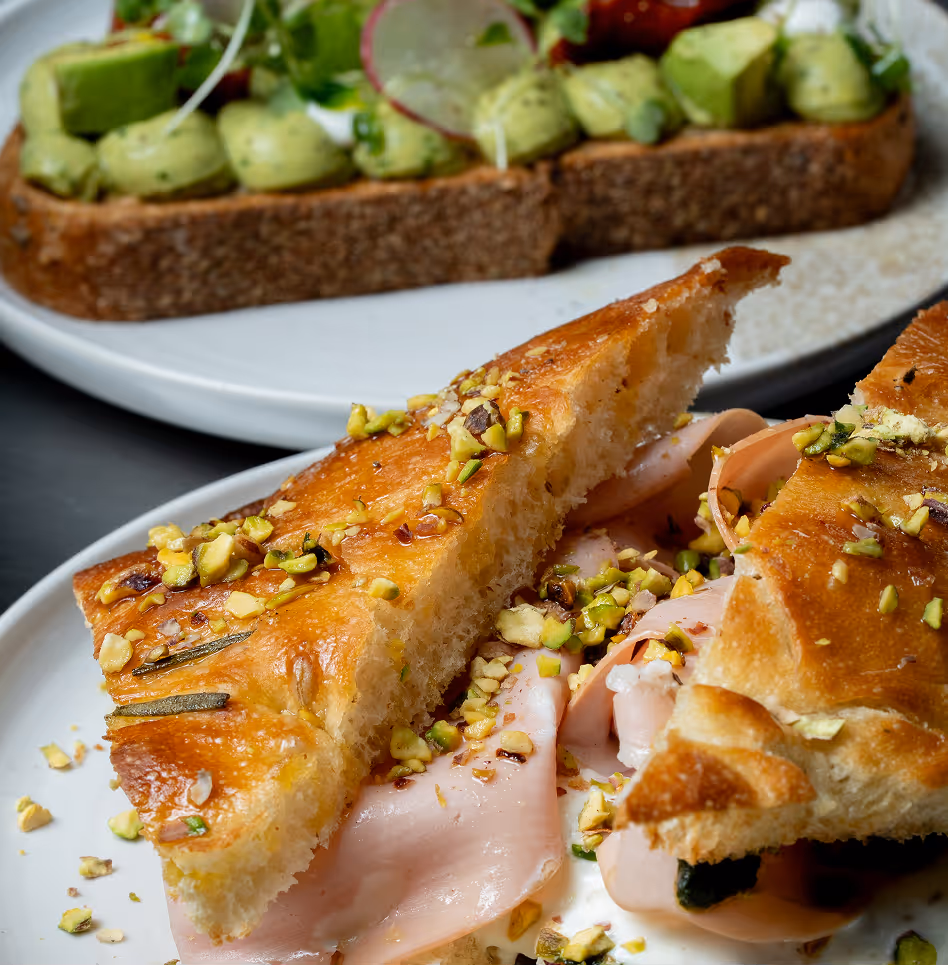 Close-up of a sandwich with focaccia bread, prosciutto, and chopped pistachios on a white plate.