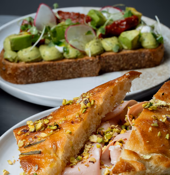 Two plates with an avocado toast topped with radishes and a sandwich with turkey and pistachio nuts.